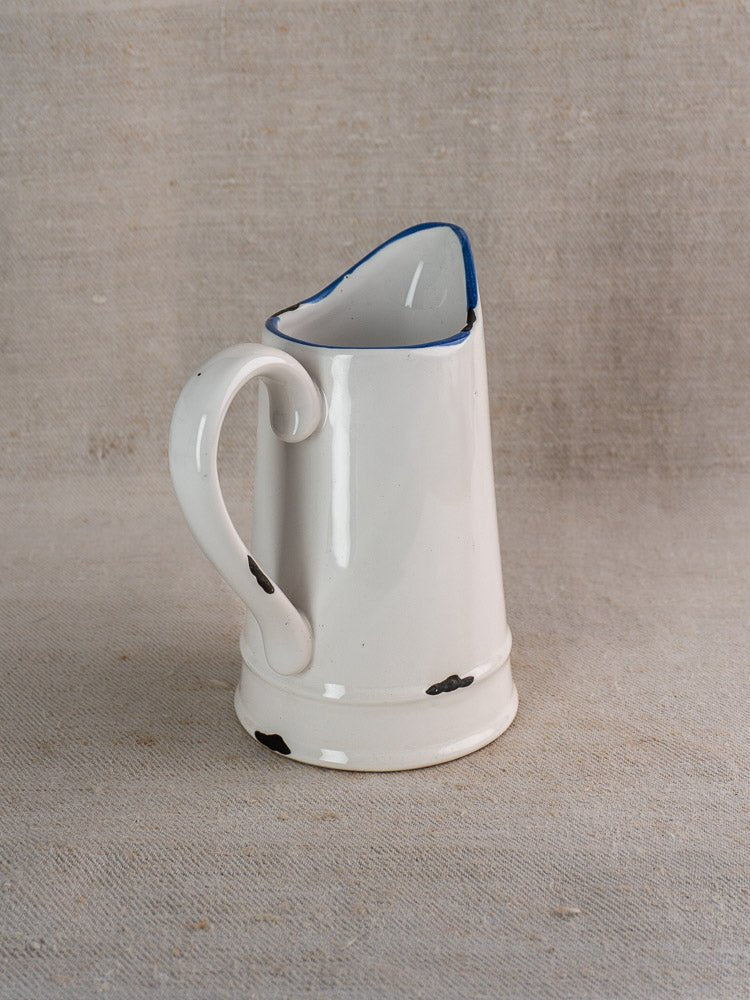 Vintage French faience pitcher with white glaze and blue trim on beige fabric surface.