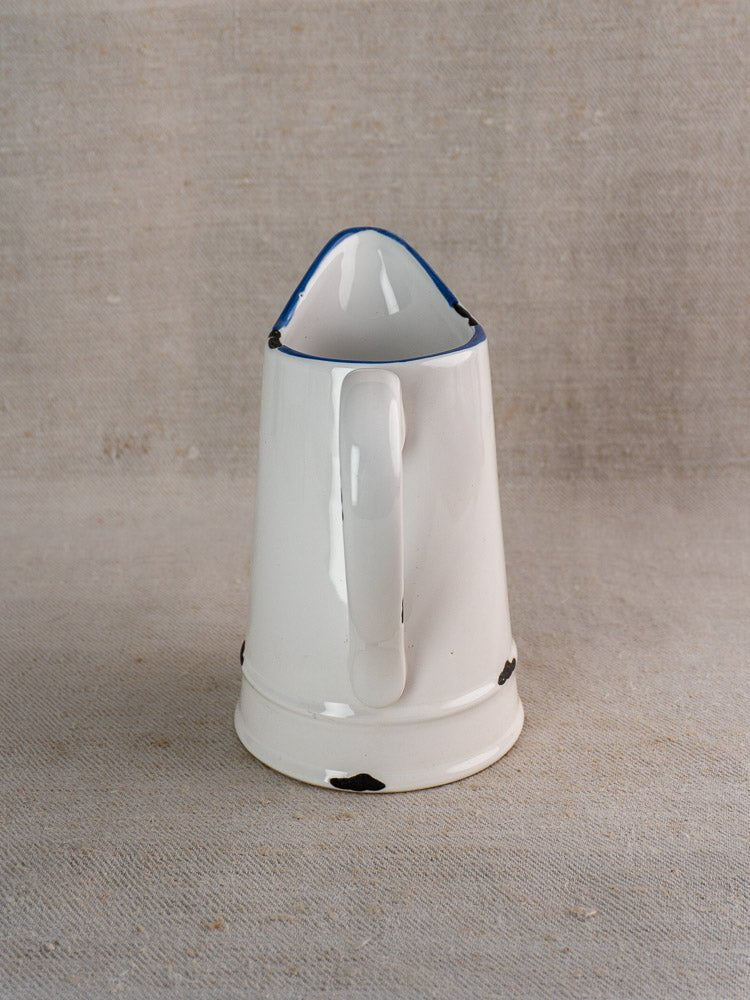 Vintage French faience pitcher with white glaze, blue trim, black chips on beige textured fabric.
