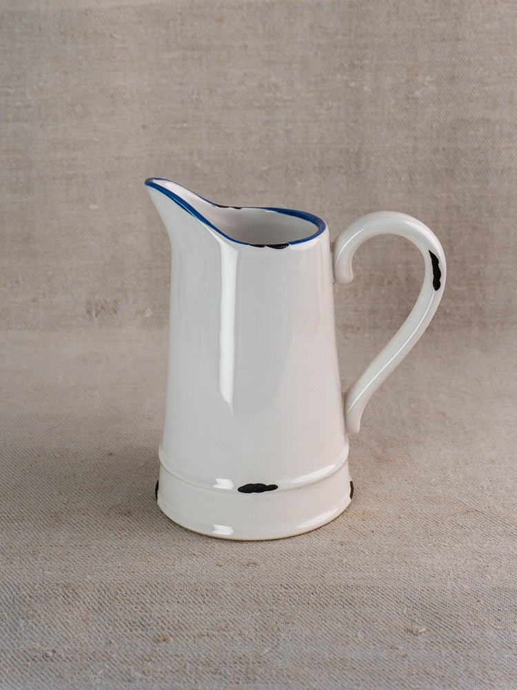 Vintage French faience pitcher with white glaze, blue rim, and distressed black spots on beige fabric.