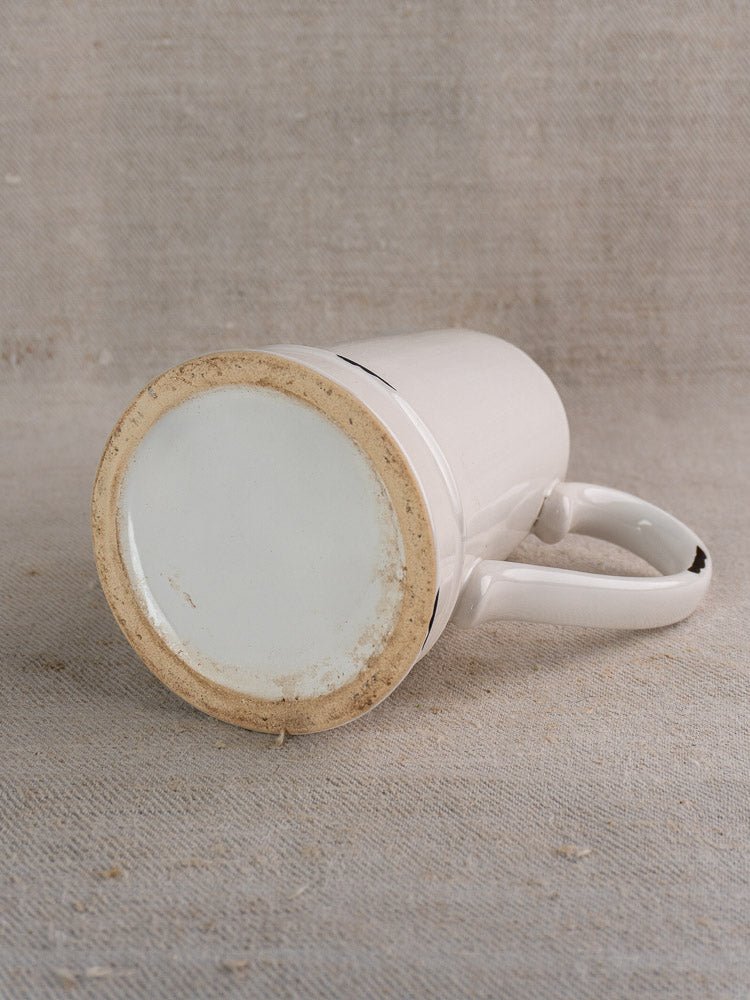 Vintage French faience pitcher with white glaze, blue trim, and visible handle on beige fabric background.