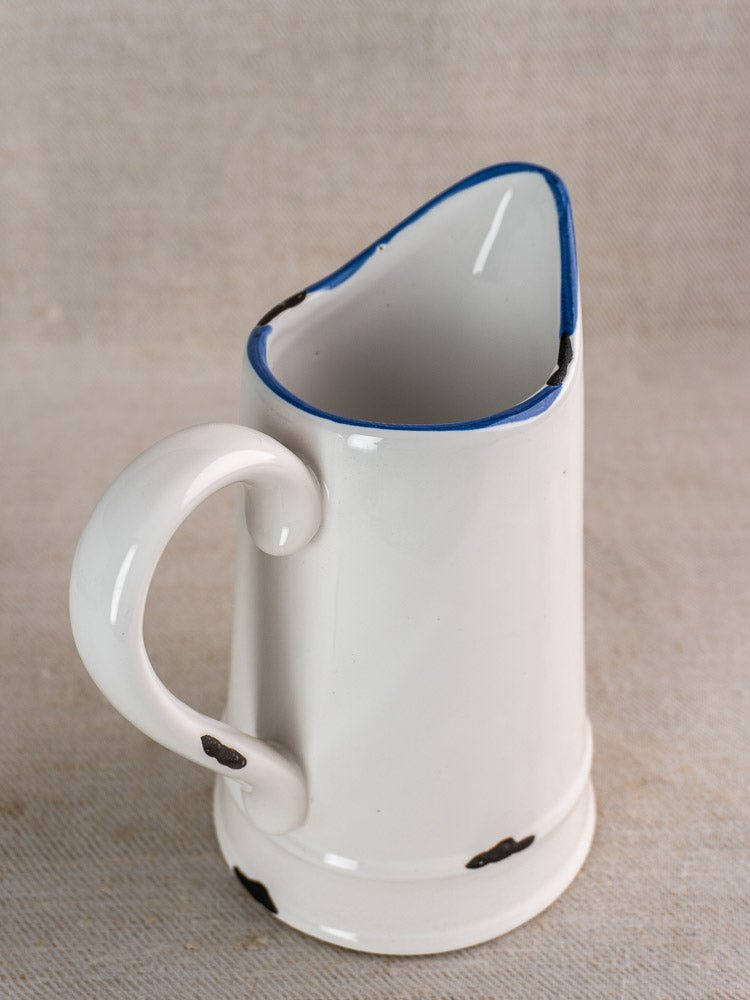 Vintage French faience pitcher with white glaze, blue rim, black spots, and large handle on beige surface.
