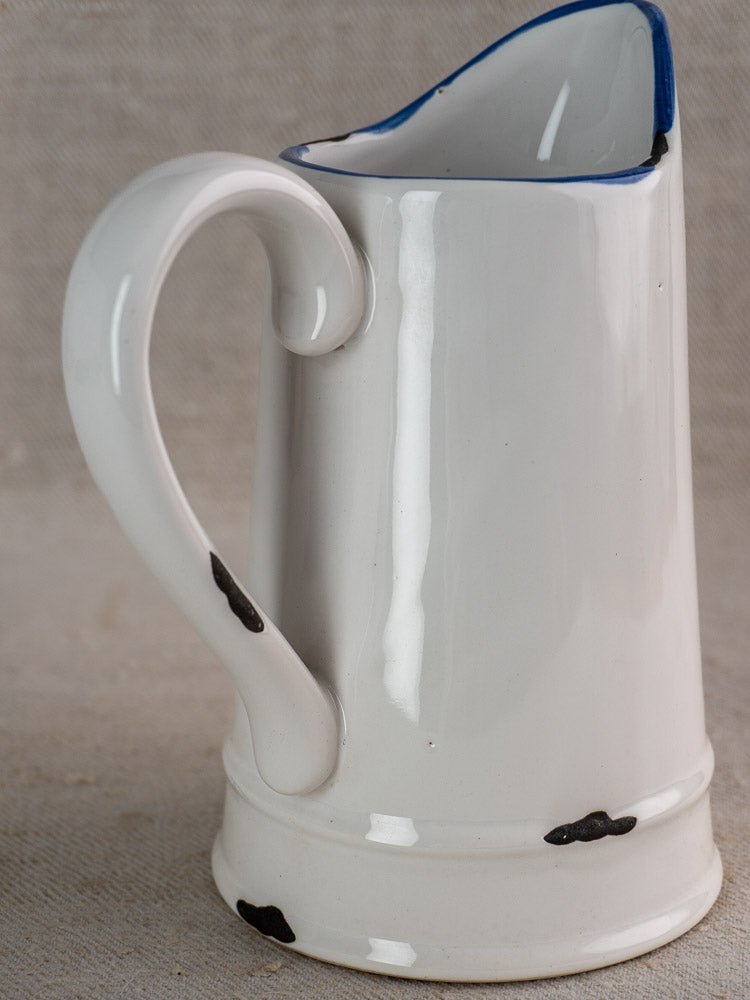 Vintage French faience pitcher with white glaze, blue trim, black spots, and curved handle.