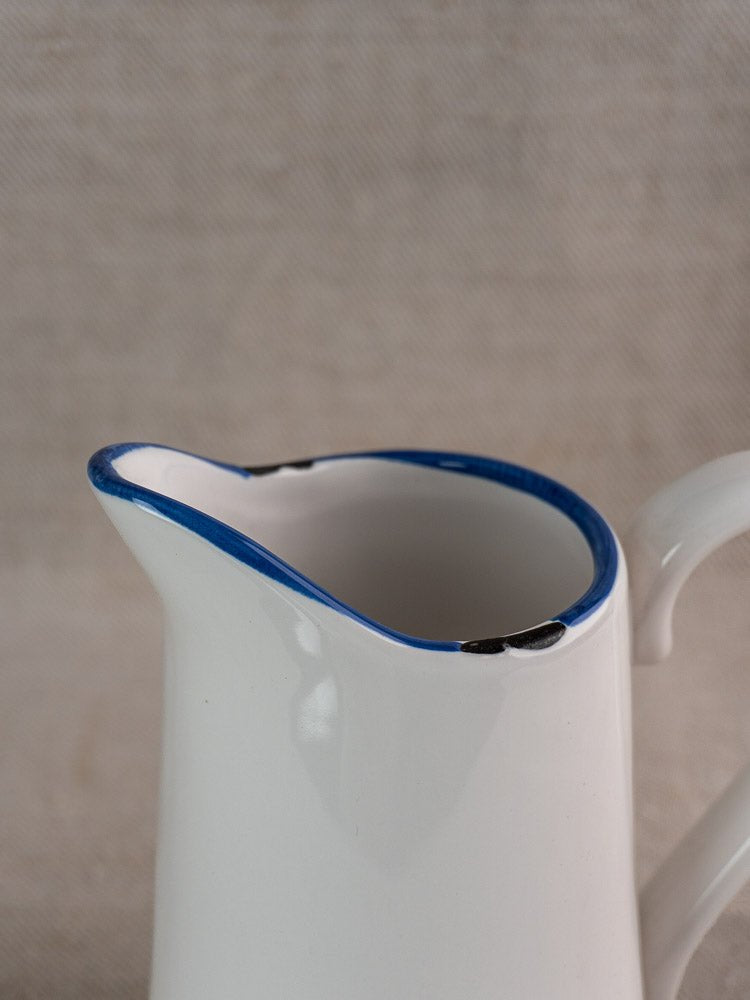 Vintage French faience pitcher with white glaze, blue trim, and gently worn edges on neutral background.