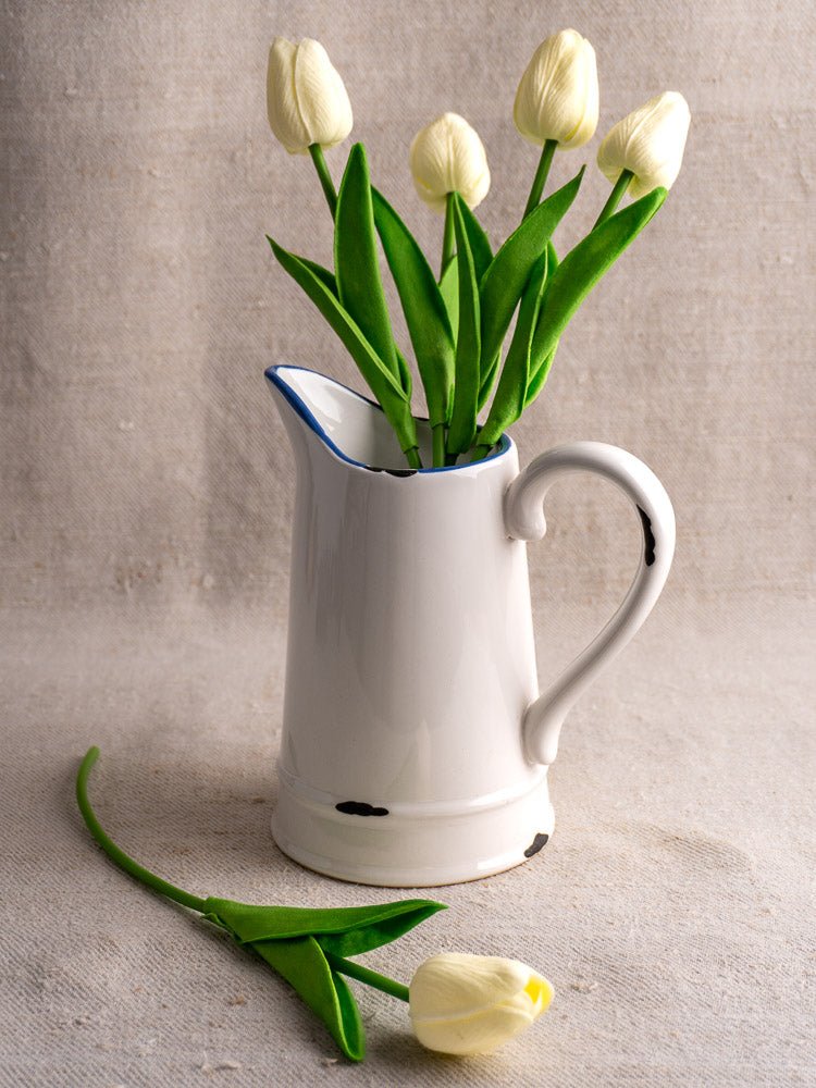 French faience pitcher with white glaze and blue trim, holding five white tulips, on beige surface.