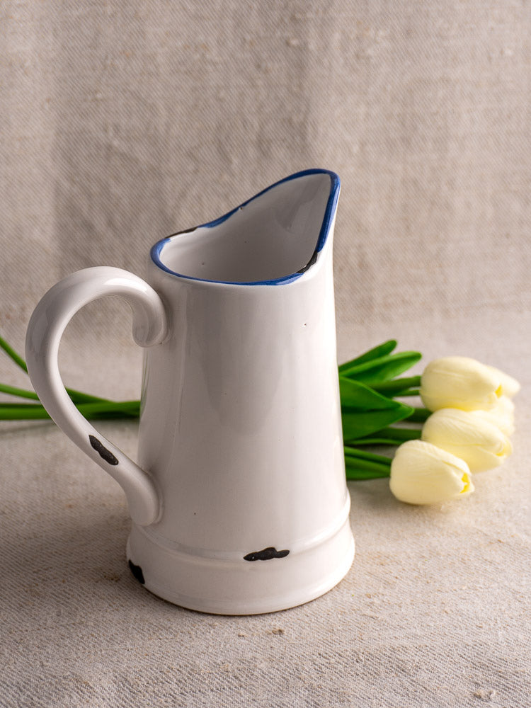 French faience pitcher with white glaze, blue rim, large handle, and rustic black specks on beige fabric.
