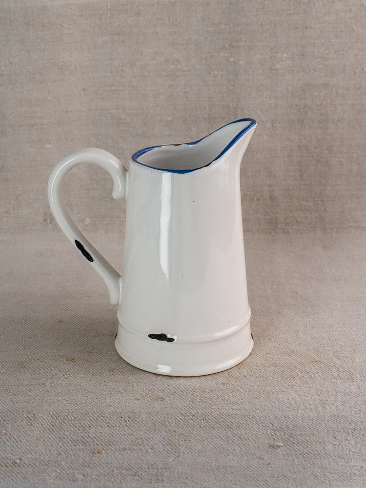 French faience pitcher with white glaze, blue rim, large handle, and rustic black specks on beige fabric.