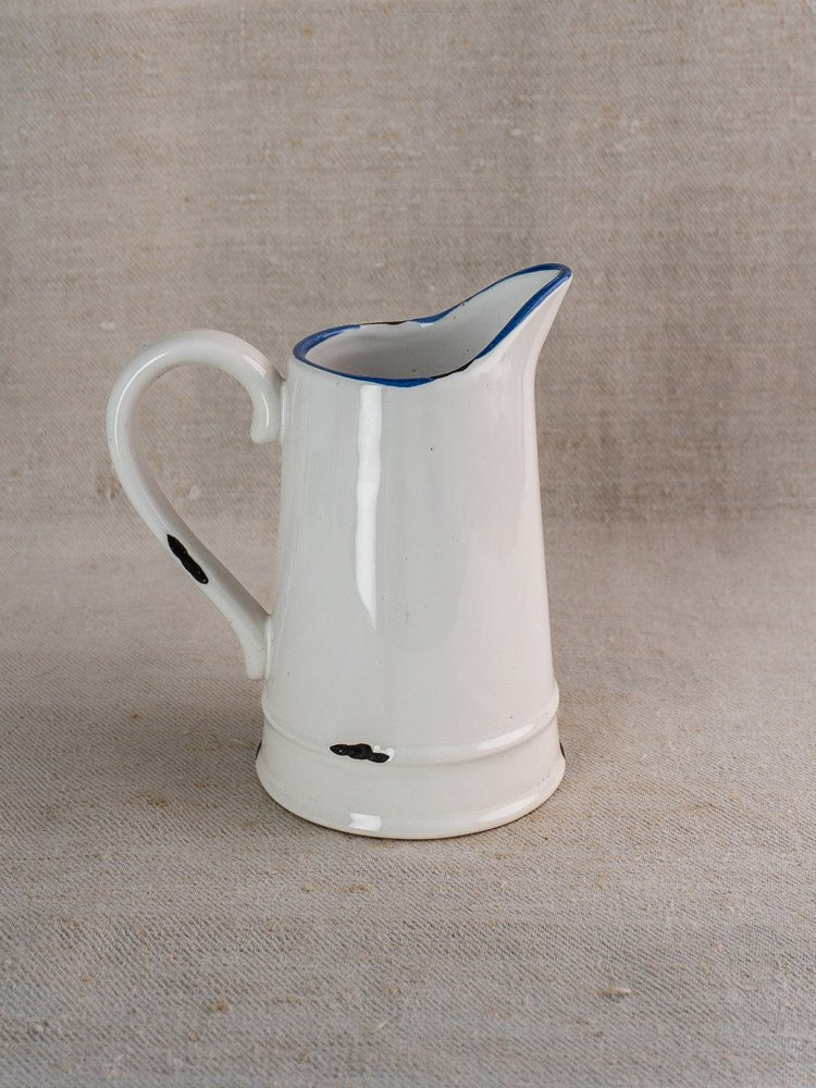 French faience pitcher with white glaze, blue rim, large handle, and rustic black specks on beige fabric.