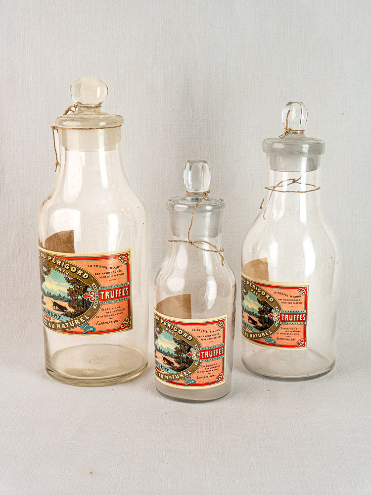 Set of three antique French L. Jaubert truffle jars with glass stoppers and classic Truffes labels on light fabric background.