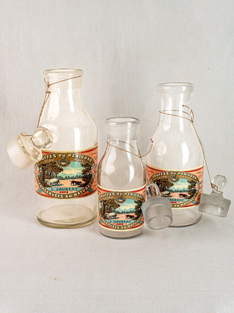 Set of three antique French L. Jaubert truffle jars with glass stoppers, landscape labels, plain backdrop.
