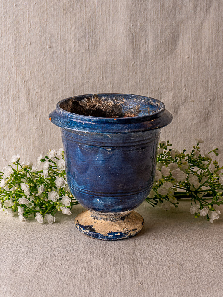 Antique French Medici blue glazed terracotta vase on a beige surface with white flowers and neutral fabric backdrop.