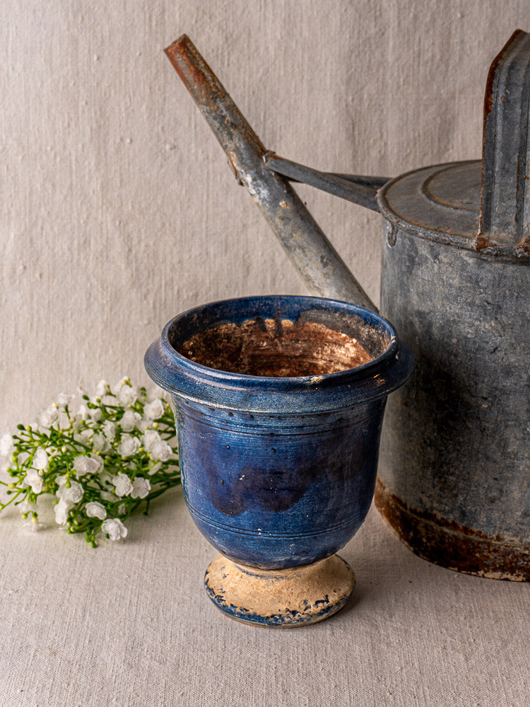 Antique French Medici Vase in blue glazed terracotta beside metal watering can, white flowers on beige fabric.