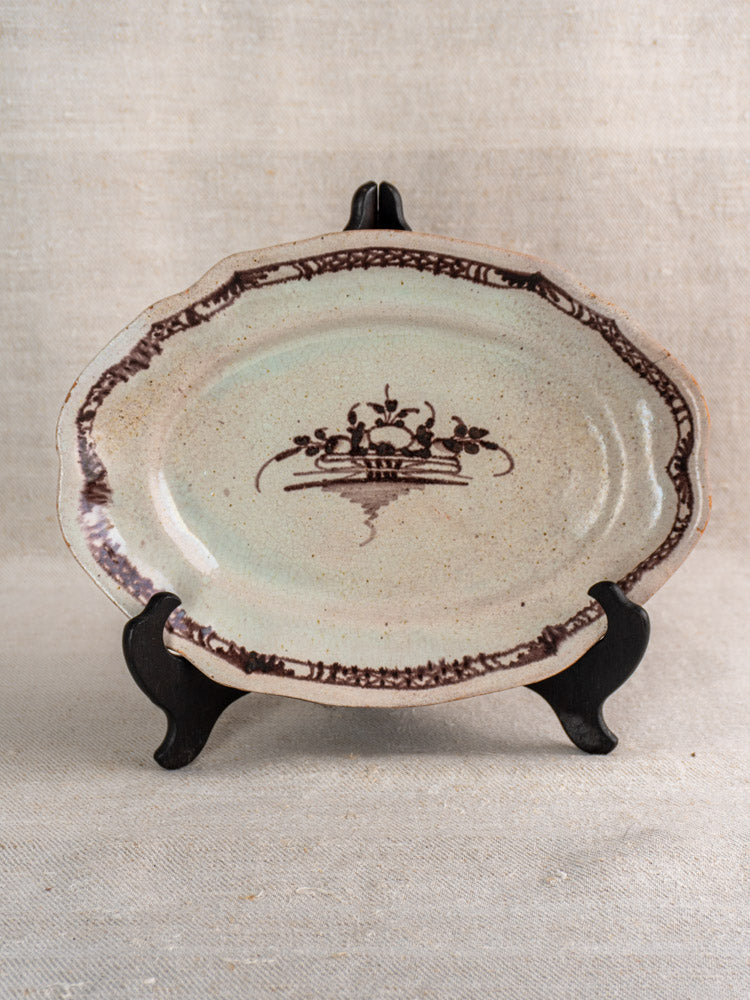 Antique French Cul Noir faience platter with brown manganese design, displayed on a black stand against beige fabric.