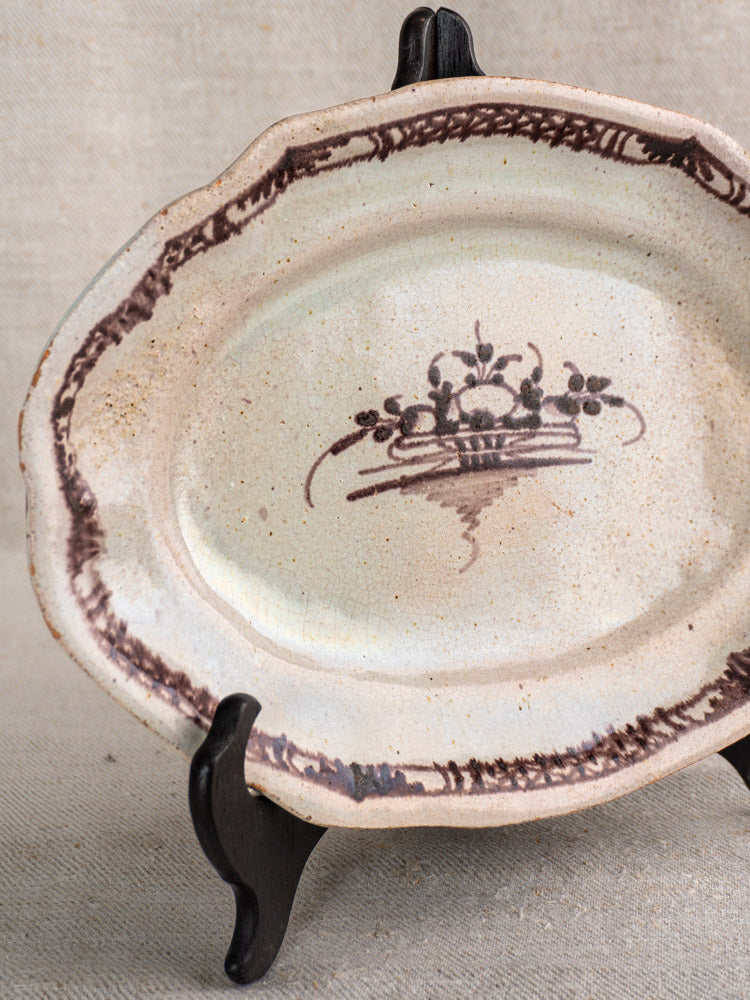 Antique French Cul Noir faience platter with brown border and fruit basket center on black stand, neutral fabric backdrop.