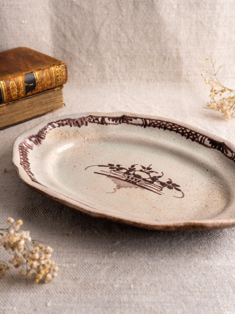 Antique French Cul Noir faience platter with brown crown motif on beige cloth beside vintage books and dried flowers.