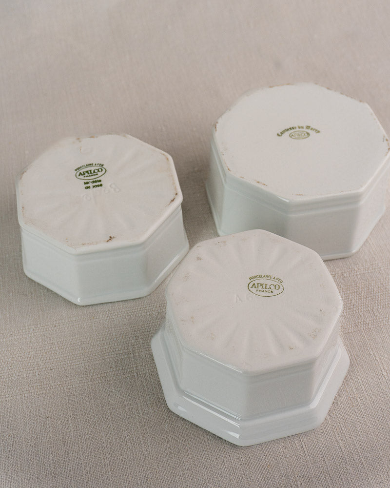 Three white Limoges porcelain Apilco octagonal dishes upside down, showing maker's mark and rim wear on fabric.