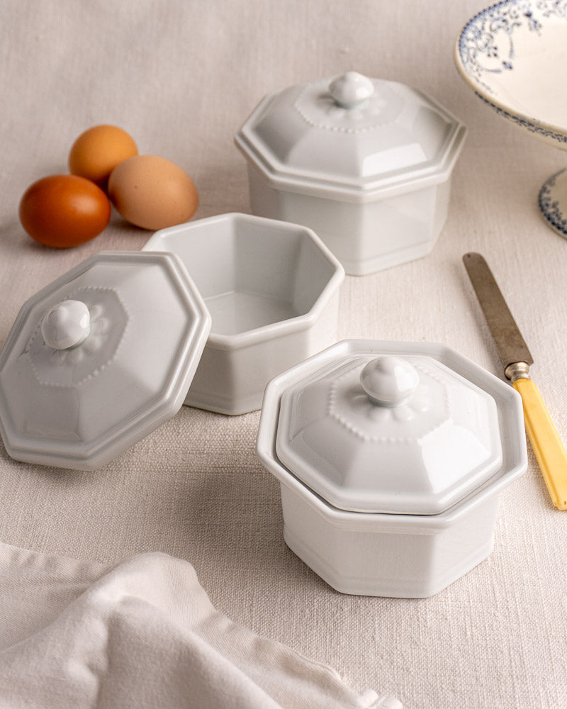 Set of three white octagonal Apilco French porcelain pots with lids on light cloth, one open.