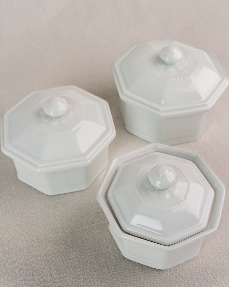 Set of three vintage white Apilco porcelain Modèle de José octagonal pots with lids on beige fabric.