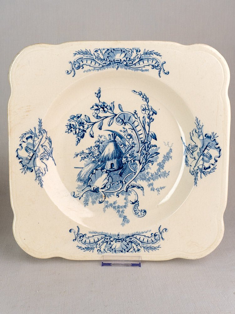 Square cream plate with ornate blue floral motifs, central pastoral scene, and Louis XV border flourishes.