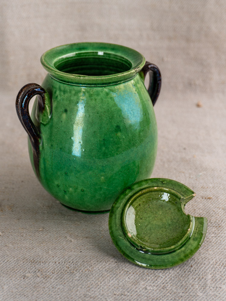 Antique French Dieulefit green earthenware olive pot with dark brown handles, notched lid, on beige fabric. Early 20th century.