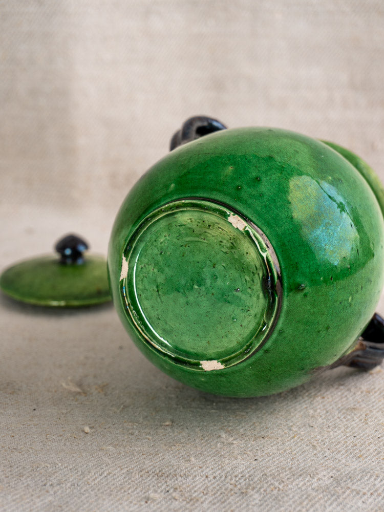 Early 20th-century French olive pot in Dieulefit green earthenware, base up, lid beside on beige textured fabric.