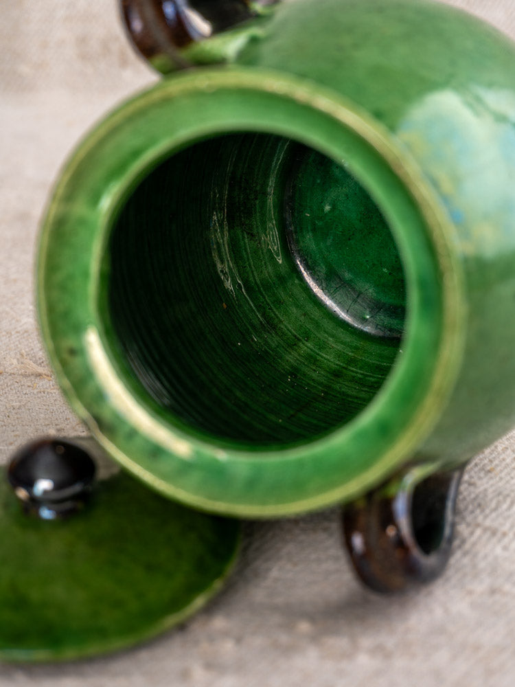 Antique French Dieulefit green earthenware olive pot with dark handles, lid removed on textured beige surface.