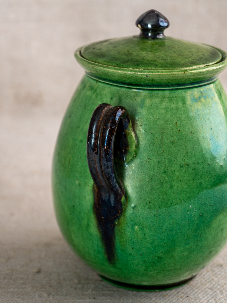 Antique French Dieulefit green earthenware olive pot with lid and curved handle on beige fabric. Glossy finish.