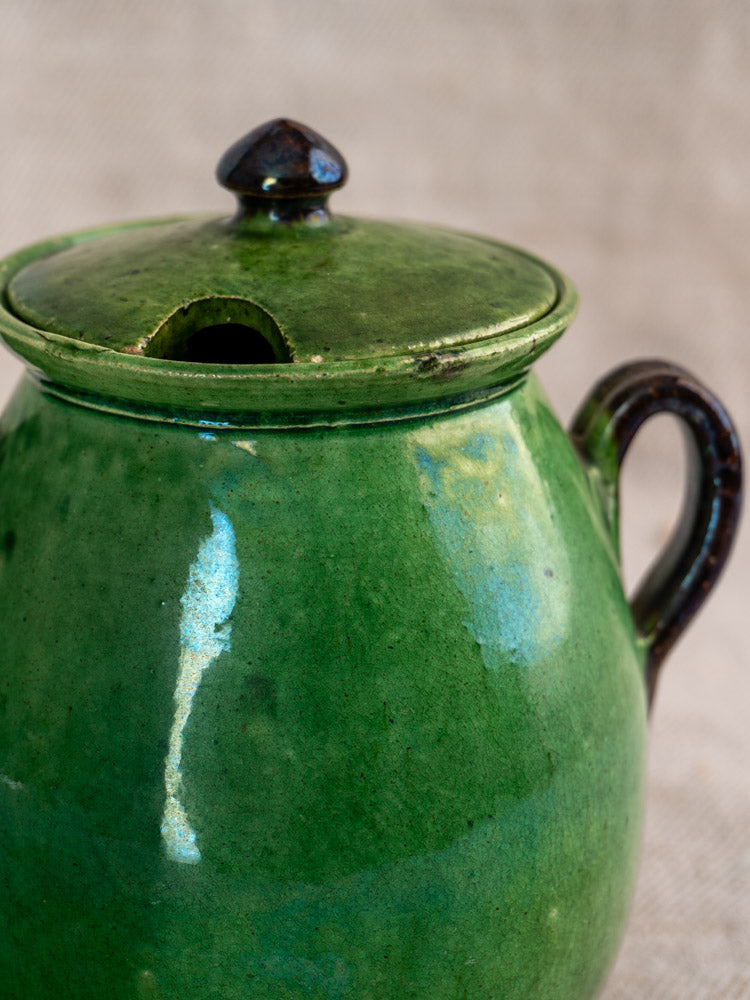Antique French Dieulefit green olive pot with round side handle and lidded notched top on beige textured background.