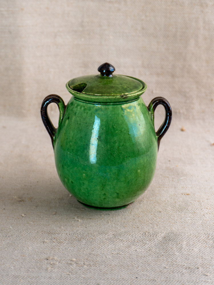 Antique French Dieulefit green earthenware olive pot with two handles and lid on beige textured fabric.