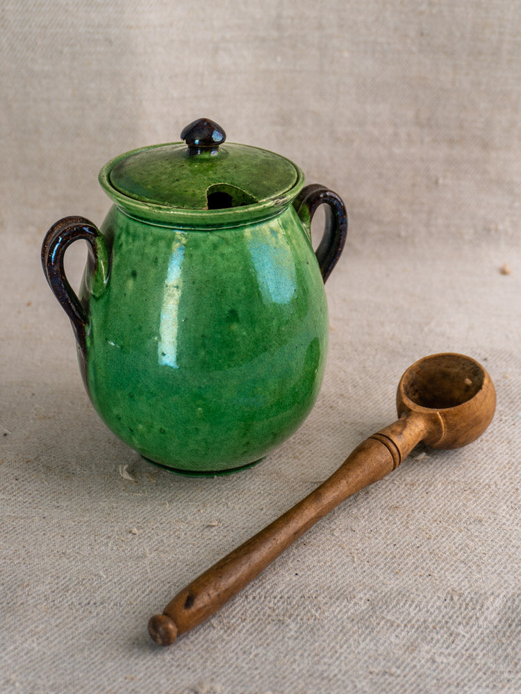 Antique French Dieulefit green earthenware olive pot with two handles and lid, on beige fabric next to a wooden ladle.
