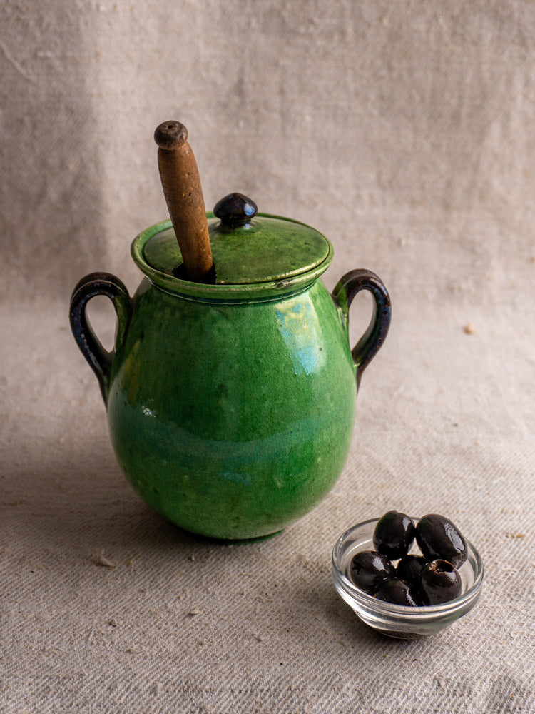 Antique French Dieulefit green earthenware olive pot with lid, wooden spoon, and handles beside black olives on beige fabric.