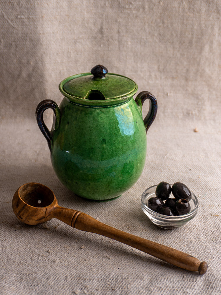 Antique French Dieulefit green earthenware olive pot with lid, on linen beside black olives and wooden ladle.