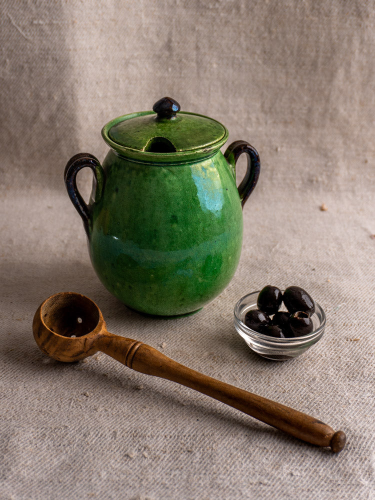 Antique French Dieulefit green earthenware olive pot with lid, two handles, black olives, and wooden ladle on beige fabric.