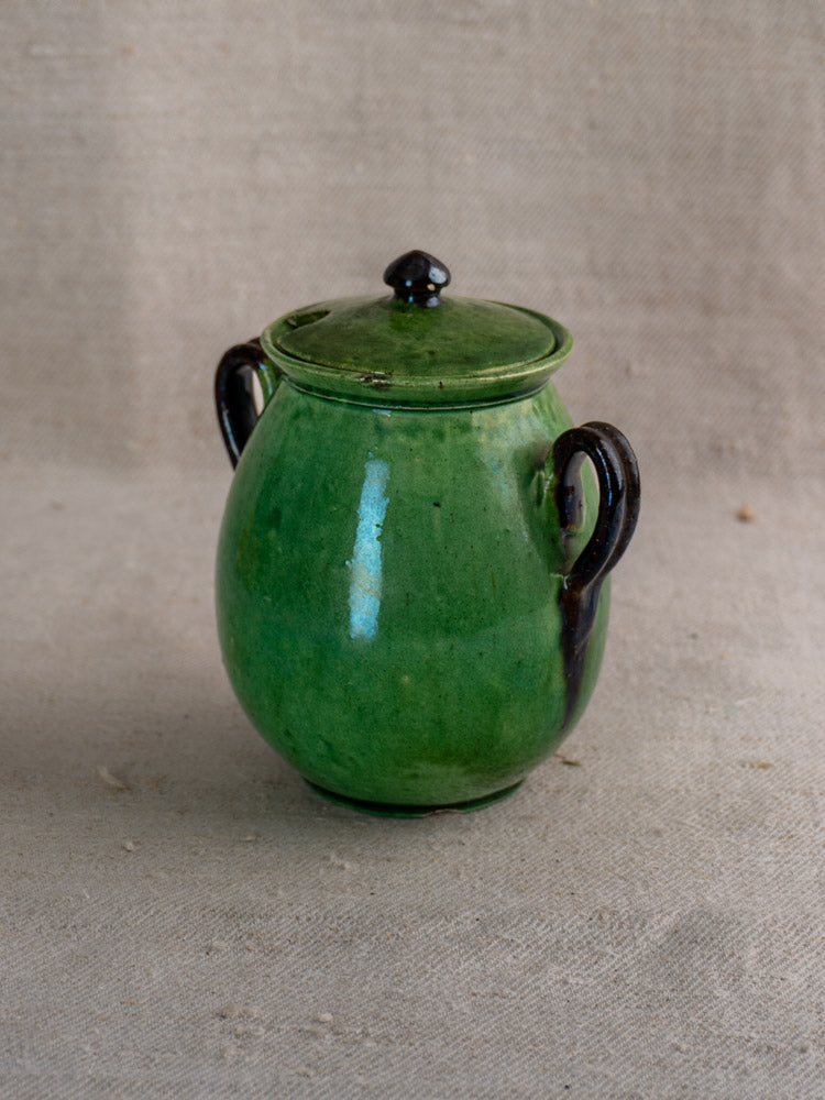 Antique French Dieulefit green earthenware olive pot with glossy finish, lid, and dark handles on beige textured fabric.