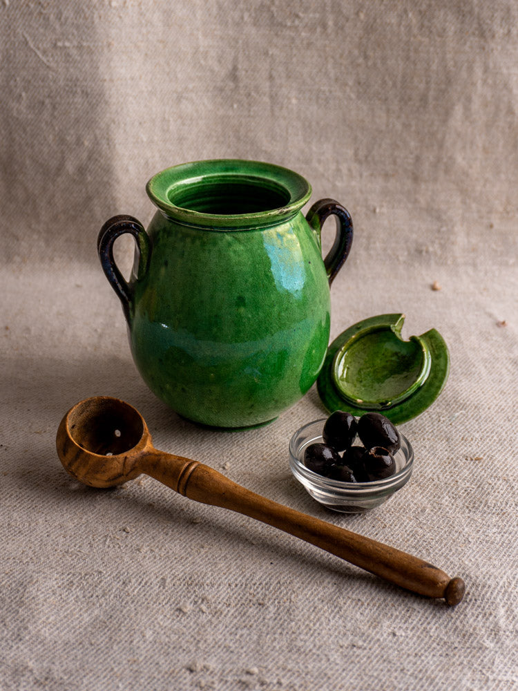 Antique French Dieulefit green earthenware olive pot with handles and lid, on linen beside spoon and bowl of olives.