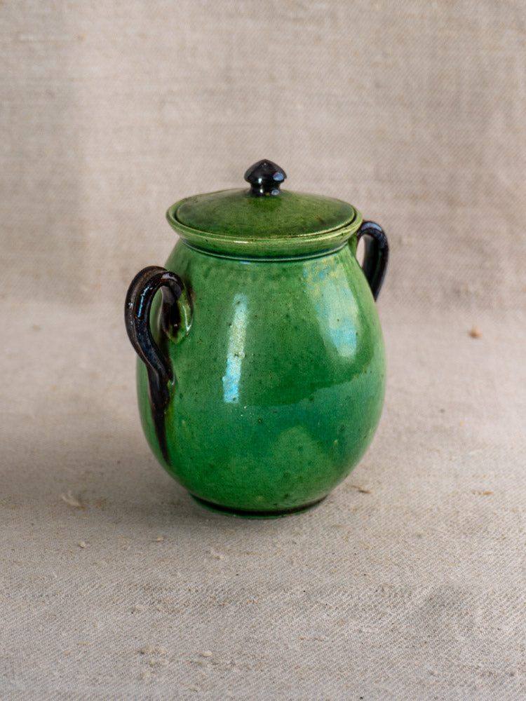 Antique French Dieulefit green earthenware olive pot with lid and black handles on beige textured fabric.