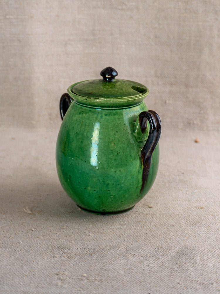 Antique French Dieulefit green earthenware olive pot with lid and dark handles on beige textured fabric background.