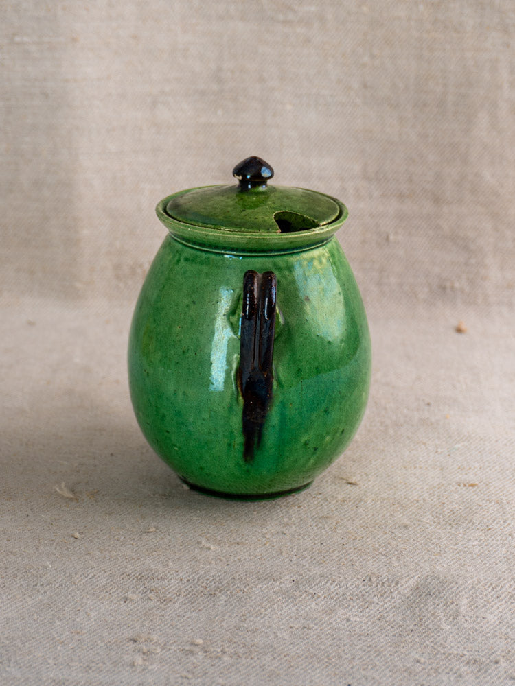 Antique French Dieulefit green earthenware olive pot with lid and black handle on textured beige fabric.