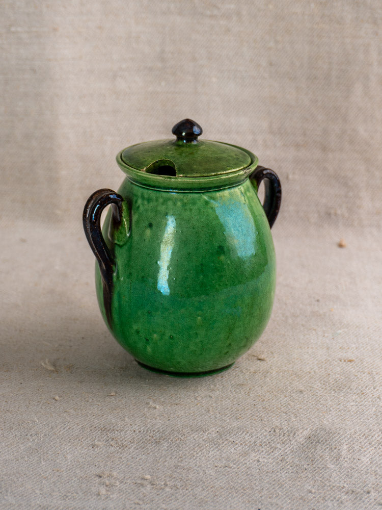 Antique French Dieulefit green earthenware olive pot with lid and dark brown handles on beige textured fabric.