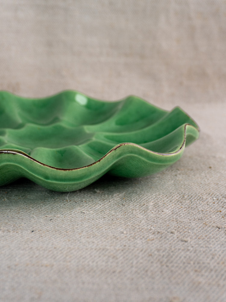 Antique French Dieulefit ceramic melon dish with emerald green glaze and wavy edges on textured beige fabric.