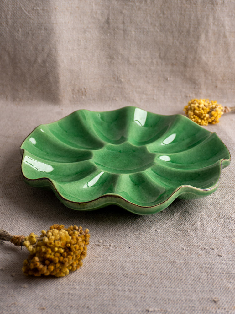 Antique French ceramic melon dish with emerald green glaze and scalloped edges on beige fabric with yellow dried flowers.
