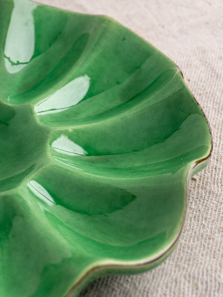 Glossy emerald green French ceramic melon dish, leaf-shaped with defined veins, on beige textured fabric.