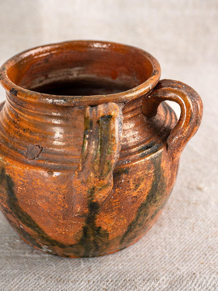 Antique French rillettes pot, rustic brown earthenware with Tigré glaze, green and black patina, on beige textured fabric.