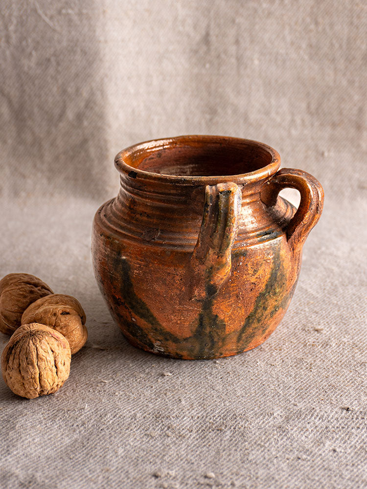 Antique French glazed earthenware rillettes pot with tigré drip glaze and handle, displayed on beige textured fabric with walnuts.
