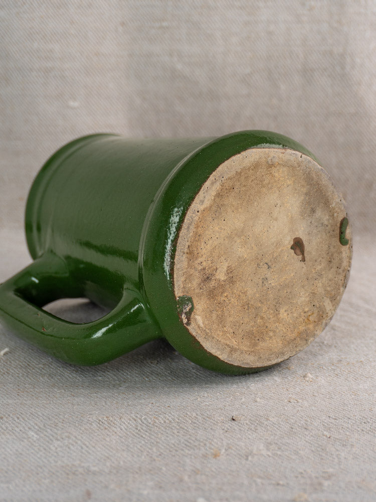 Antique French emerald green earthenware pitcher, unglazed base visible, on textured beige fabric background.