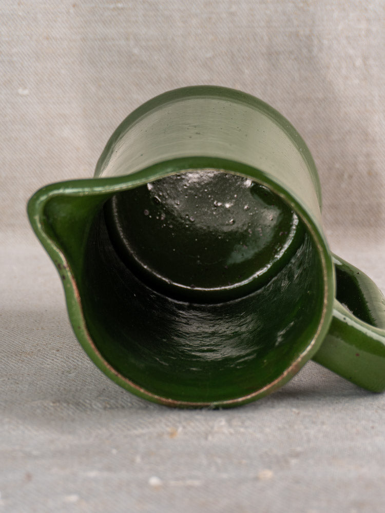 Antique French emerald green earthenware pitcher displays interior and spout on a beige textured surface.