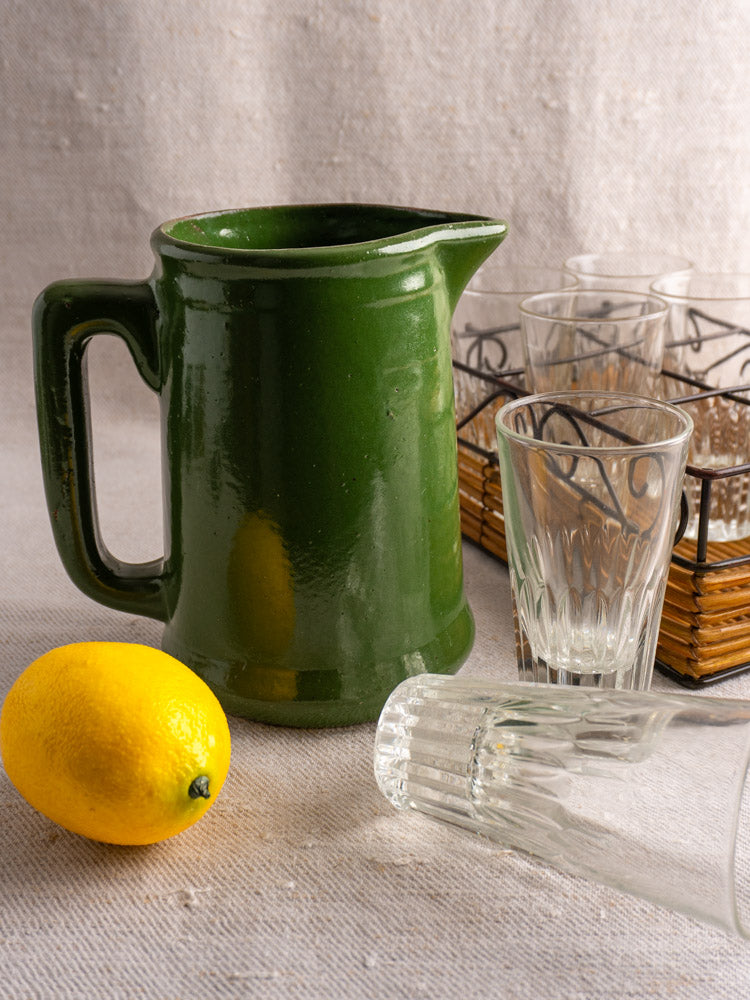 Antique French emerald green glazed earthenware pitcher with lemon and clear glasses on light fabric background.