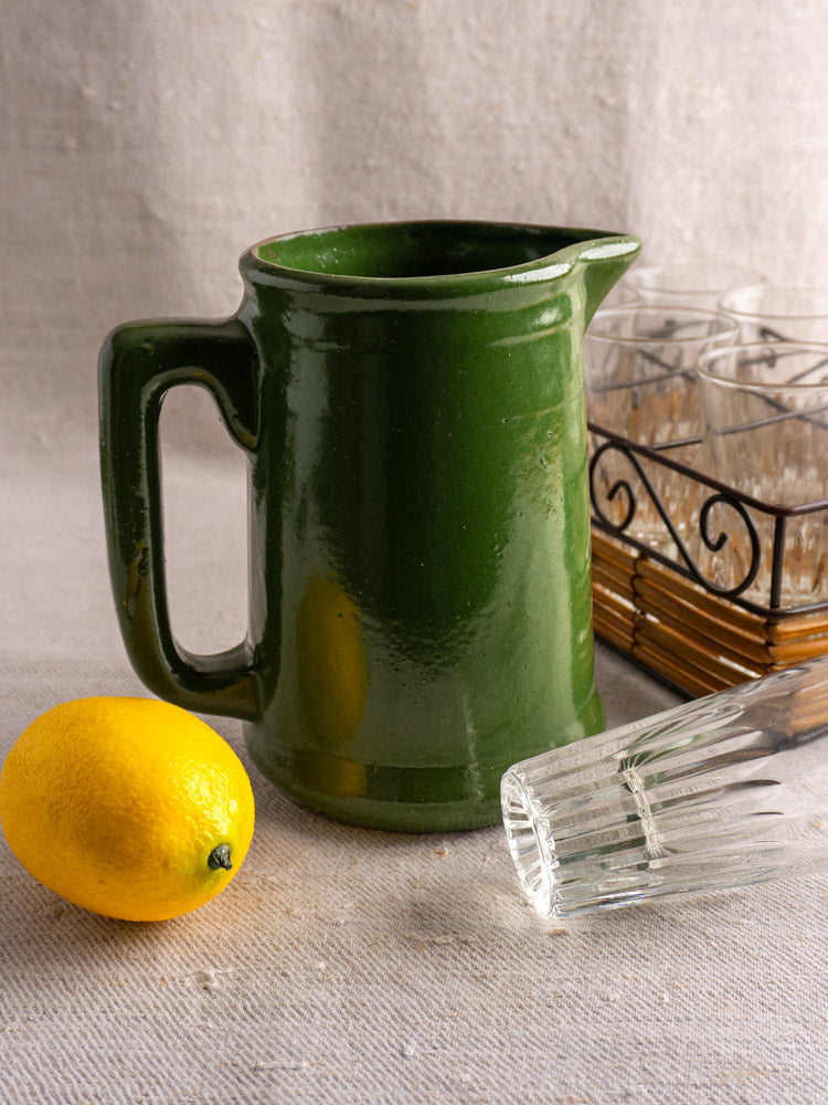 Antique French emerald green earthenware pitcher on beige fabric with lemon, glass, and metal tray of upright glasses.