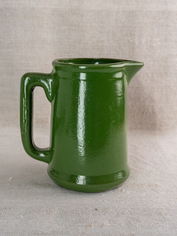Antique French emerald green glazed earthenware pitcher with large handle and spout on beige fabric background.