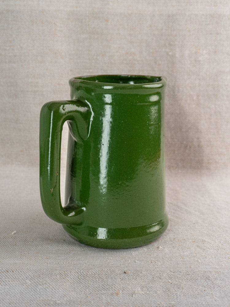 Antique French emerald green glazed earthenware pitcher on a light beige textured surface with neutral fabric backdrop.