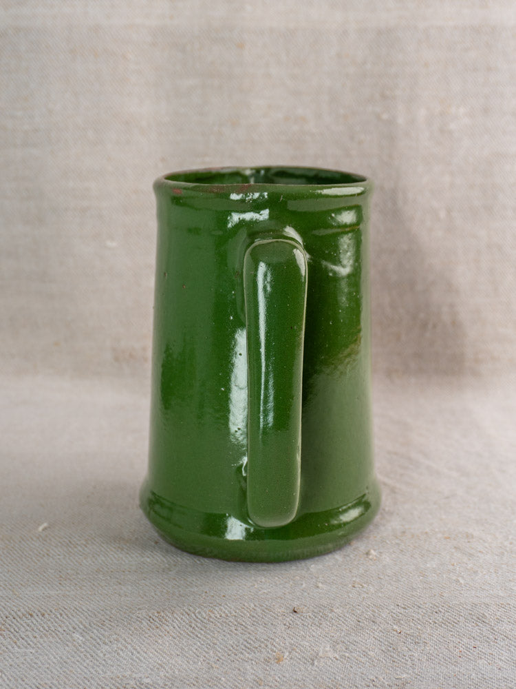 Antique French emerald green glazed earthenware pitcher, handle forward, on light beige fabric surface. Early to mid 20th century.